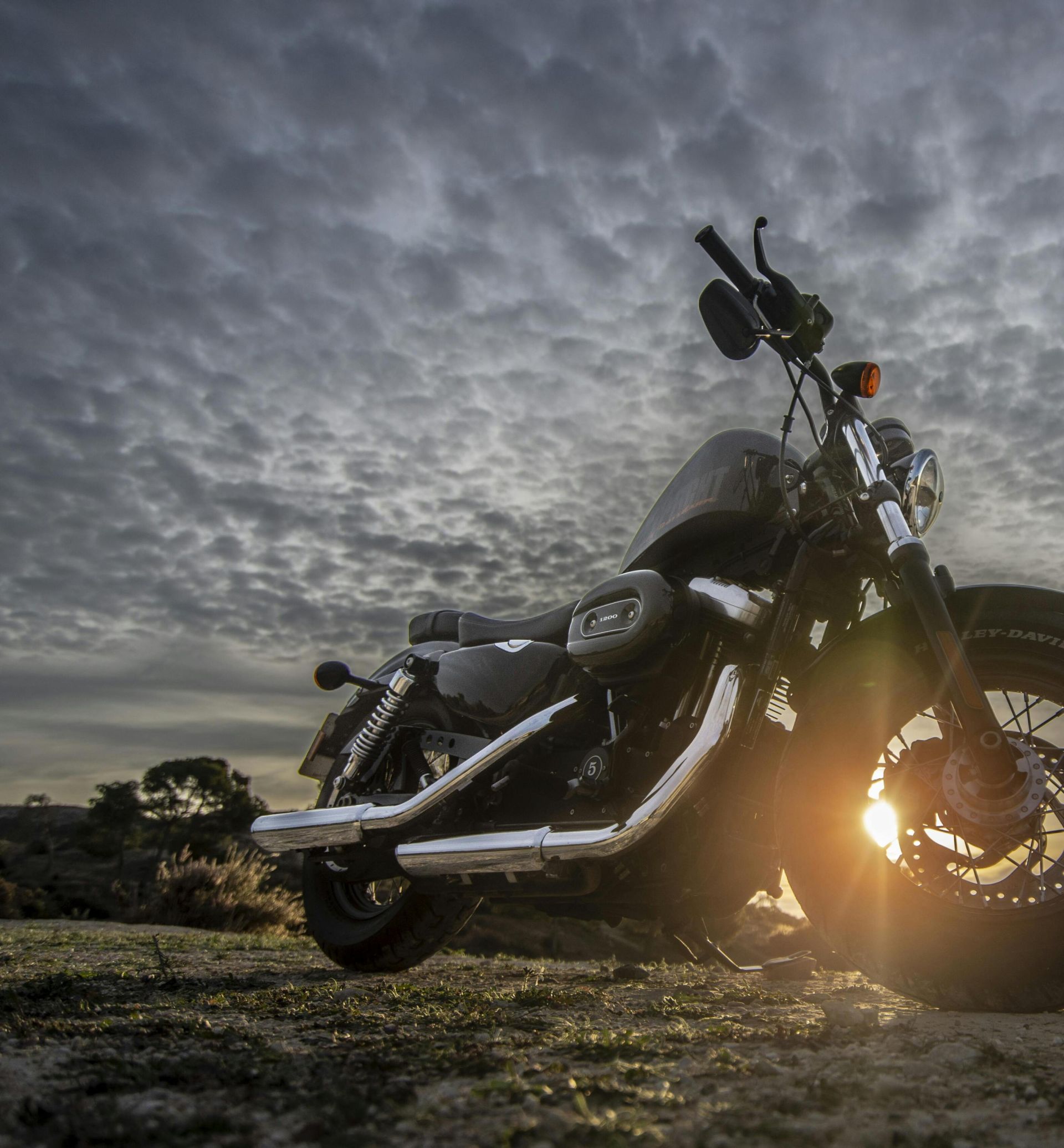 A striking image of a motorcycle parked on a dirt road with a dramatic sky at sunset, capturing the sun glare.