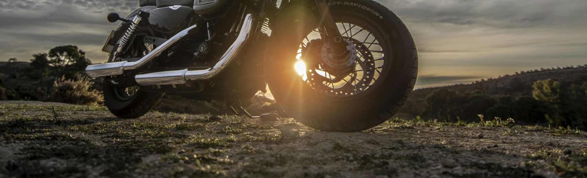 A striking image of a motorcycle parked on a dirt road with a dramatic sky at sunset, capturing the sun glare.