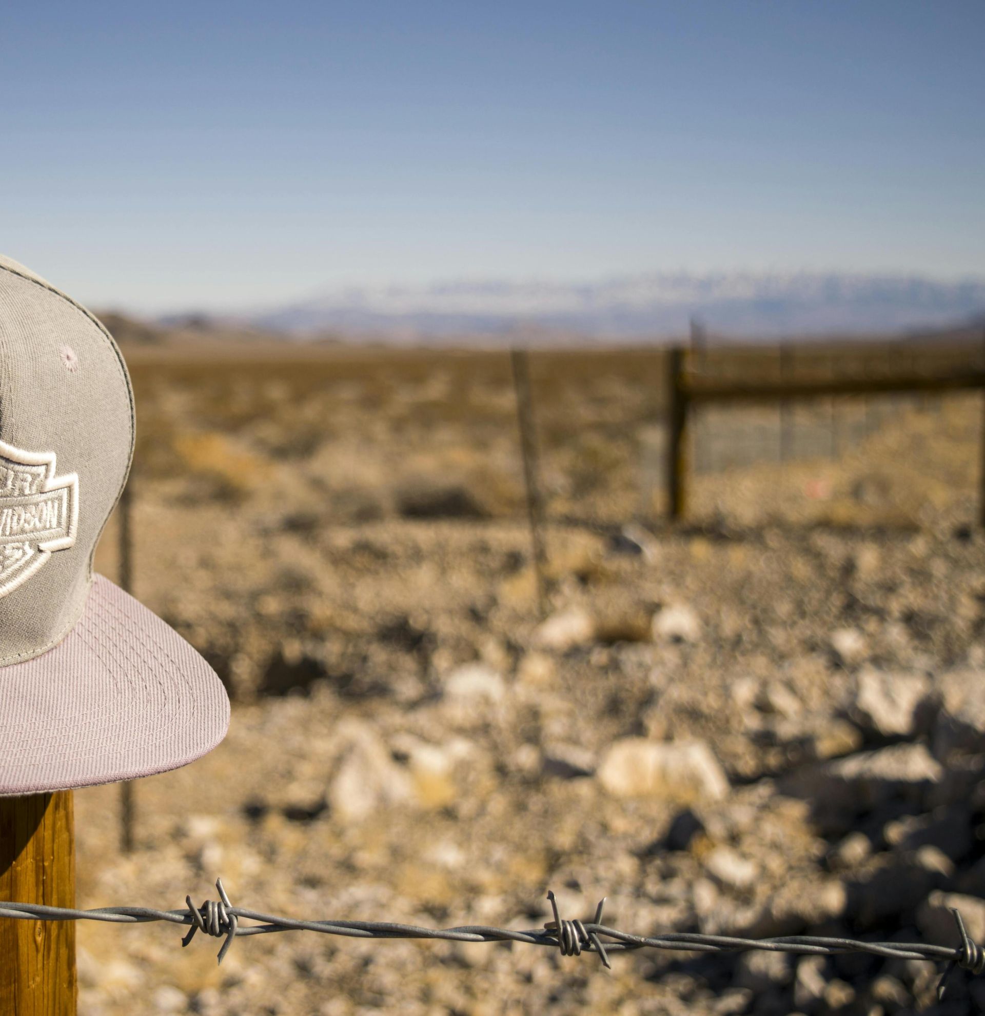 A Harley Davidson cap sits on a fence post in an arid desert landscape, under a bright blue sky.